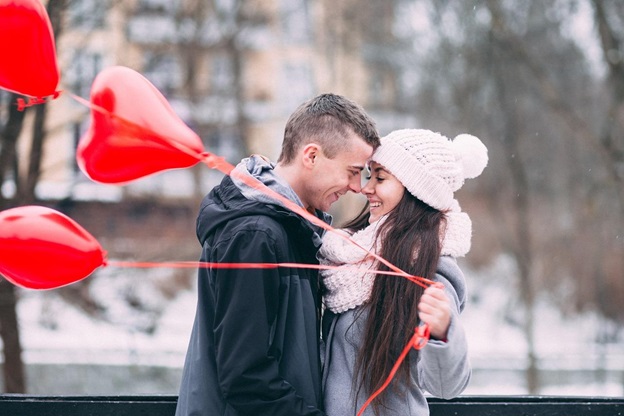 A man and a woman standing in a park during winter, and the woman is holding three red heart balloons